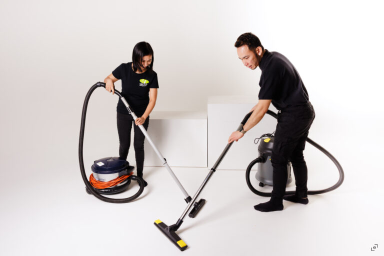 Kaksi siivoojaa imuroimassa lattiaa ammattikäyttöön tarkoitetuilla imureilla valkoisella taustalla